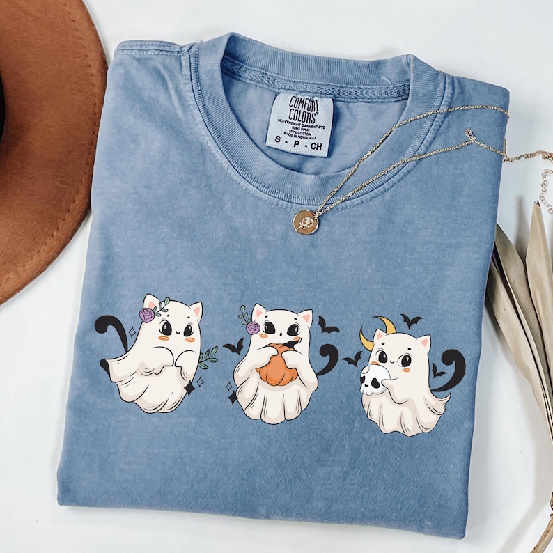 May include: A blue Comfort Colors t-shirt featuring three cartoon ghost cats. The ghosts have floral accents, pumpkins, and skulls. A gold necklace with a pendant is draped across the shirt. The shirt is a small size.