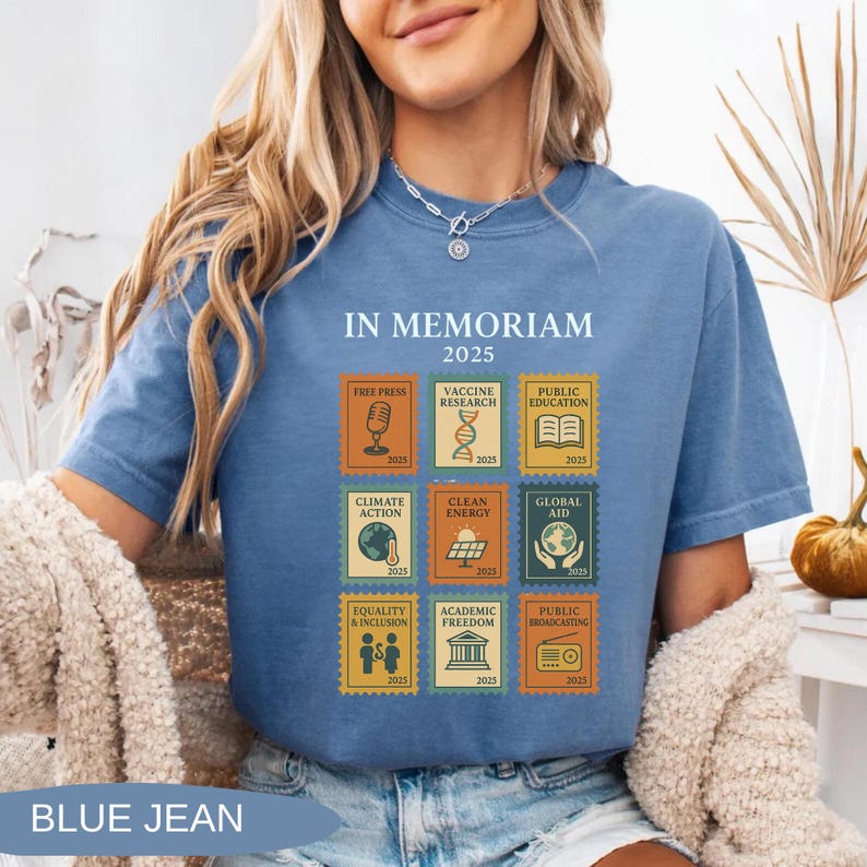 May include: Blue jean-colored t-shirt with the text "IN MEMORIAM 2025". The shirt features nine postage stamp-style graphics with themes like "Free Press", "Vaccine Research", and "Public Education". The shirt is made of a soft material.