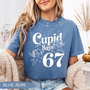 May include: Blue jean-colored t-shirt with the text "Cupid Says 67" in white. The design includes two cherubs with bows and arrows. The shirt is a short-sleeved, casual style.
