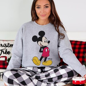 Op de afbeelding: Een grijze sweatshirt met een Mickey Mouse-afbeelding, met een rode broek en gele schoenen. Het Disney-logo is zichtbaar. De sweatshirt wordt gedragen met een zwart-wit geruite pyjamabroek.