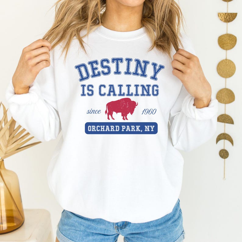 May include: White sweatshirt with the words 'DESTINY IS CALLING' in blue block letters. Below the text is a red buffalo graphic and the words 'since 1960'. Underneath is the text 'ORCHARD PARK, NY' in a blue rectangle.