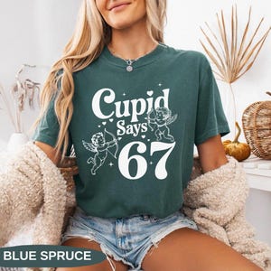 May include: A teal-colored t-shirt with the text "Cupid Says 67" in white. The design includes two cherubs with bows and arrows. The words "Blue Spruce" are at the bottom of the image.