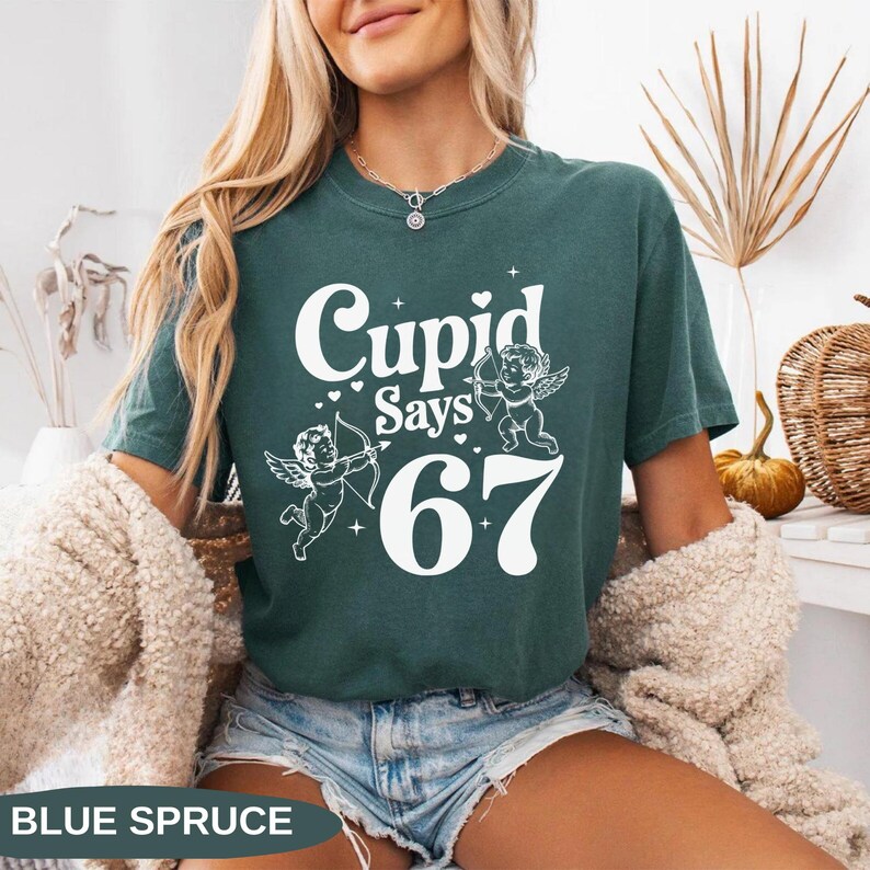 May include: A teal-colored t-shirt with the text "Cupid Says 67" in white. The design includes two cherubs with bows and arrows. The words "Blue Spruce" are at the bottom of the image.