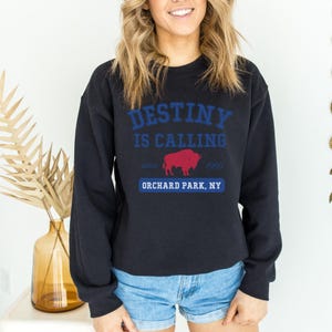May include: Black crew neck sweatshirt with the text "DESTINY IS CALLING" in blue, a red buffalo graphic, and "ORCHARD PARK, NY" in blue. The sweatshirt also has the text "since 1960".