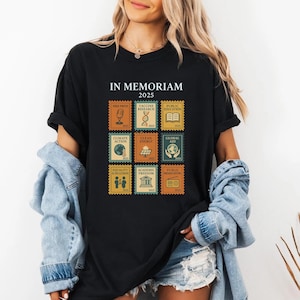 May include: Black t-shirt with the text "IN MEMORIAM 2025" and postage stamp-style graphics. The stamps feature icons and text related to social and environmental issues, including "FREE PRESS" and "CLIMATE ACTION."