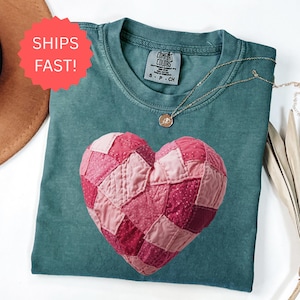 Peut inclure: Un t-shirt Comfort Colors de couleur sarcelle avec un motif de cœur en patchwork dans des tons roses. Un collier en or avec un pendentif est posé sur le t-shirt. Le t-shirt est plié et présenté sur une surface blanche.