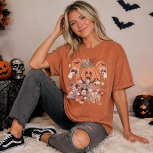 May include: A rust-colored short-sleeve t-shirt featuring a graphic print of various cartoon characters dressed in Halloween costumes, including a large central pumpkin, ghosts, and bats, paired with distressed dark gray denim jeans.
