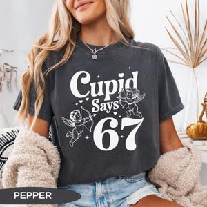 May include: Dark gray t-shirt with the text "Cupid Says 67" in white. The design includes two cherubs with bows and arrows, and small heart accents. The shirt is a casual style.