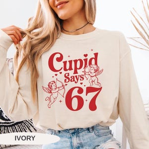 May include: Ivory-colored sweatshirt with the text "Cupid Says 67" in red. The design includes two cherubs with bows and arrows. The sweatshirt is a long-sleeved, crew neck style.