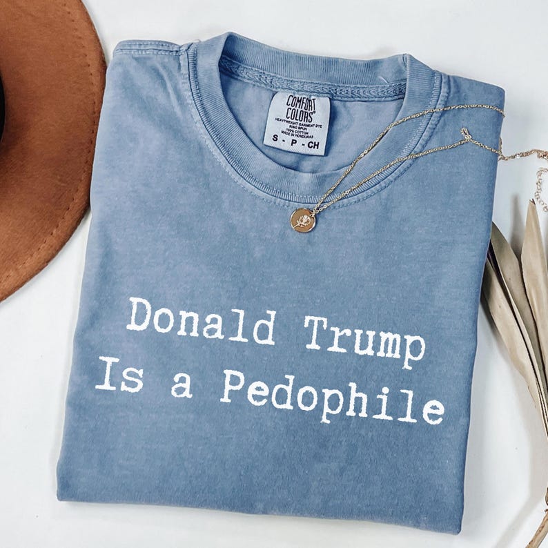 以下が含まれることがあります： 「Donald Trump Is a Pedophile」の白い文字が入った青いComfort ColorsのTシャツ。ペンダント付きのゴールドネックレスがシャツの上にかけられています。茶色の帽子が左上に一部見えています。