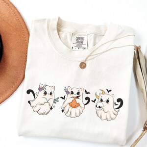 May include: Cream-colored t-shirt featuring three cartoon ghost cats with Halloween-themed accessories. The cats are adorned with floral crowns, pumpkins, and skulls. A gold necklace and a brown hat are also visible.