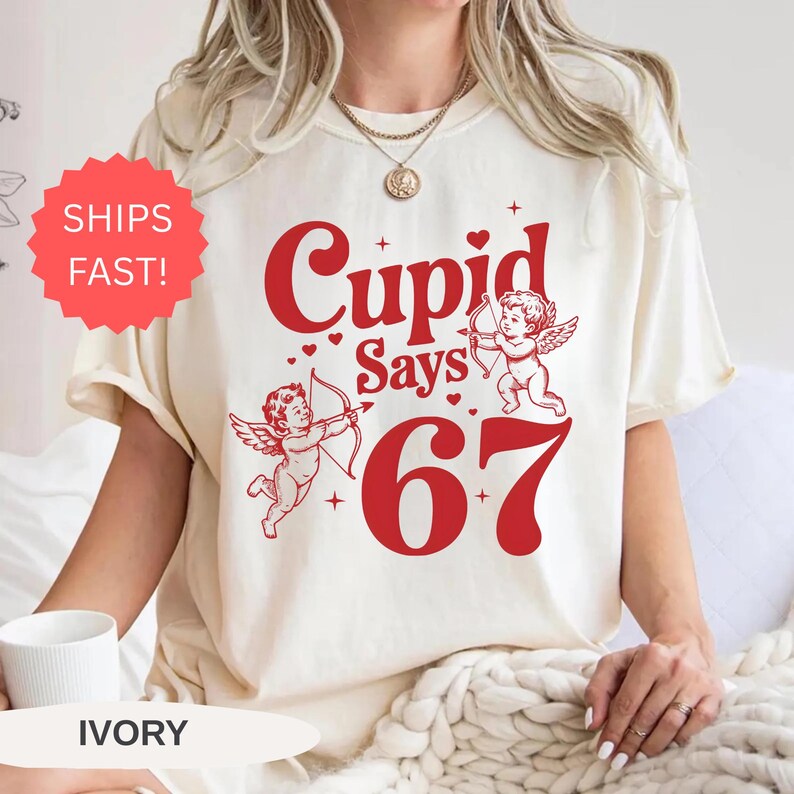 May include: Ivory-colored t-shirt with a red graphic that reads "Cupid Says 67" and features two cherubs with bows and arrows. The shirt has a crew neck and short sleeves. A red sticker that says "Ships Fast!" is visible.
