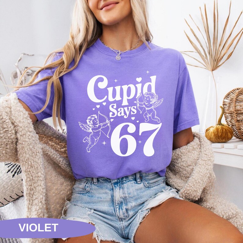 May include: A violet t-shirt with the text "Cupid Says 67" in white, along with illustrations of cupids. The shirt is made of a soft material and is suitable for casual wear.