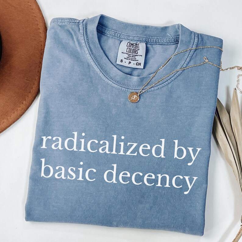 Puede incluir: Una camiseta azul Comfort Colors con la frase "radicalized by basic decency" impresa en blanco. Un collar dorado con un colgante est&aacute; drapeado sobre la camiseta. Un sombrero marr&oacute;n y hojas secas est&aacute;n en el fondo.