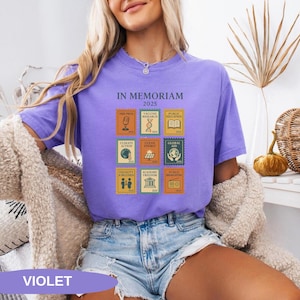 May include: Violet-colored t-shirt with the text "IN MEMORIAM 2025" and postage stamp-style graphics. The stamps depict themes like free press, vaccine research, and climate action. The word "VIOLET" is at the bottom of the image.