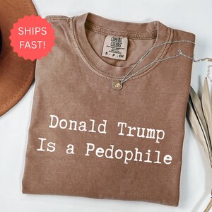 以下が含まれることがあります： 「Donald Trump Is a Pedophile」の白文字がプリントされた茶色のComfort Colors Tシャツ。小さなペンダントが付いたシルバーのネックレスがシャツの上にあります。「Ships Fast!」と書かれた赤いステッカーが左上にあります。