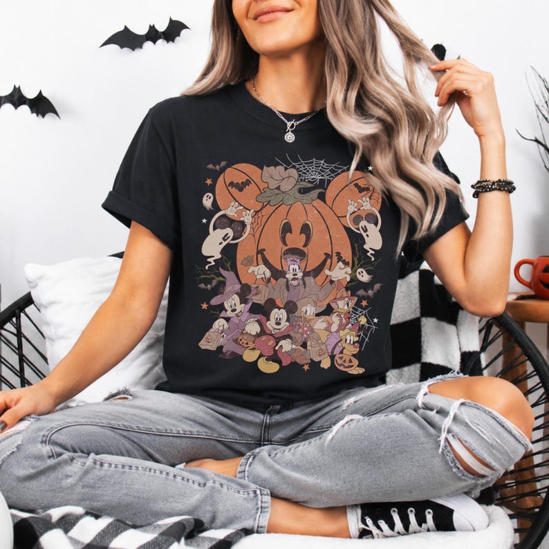 May include: A black short-sleeve t-shirt featuring a large graphic print of various cartoon characters dressed in Halloween costumes, including a central pumpkin, ghosts, and bats. The design includes orange, brown, and white colors on the black fabric.