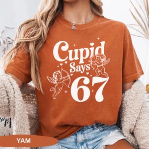 May include: Burnt orange t-shirt with the text "Cupid Says 67" in white. The design includes two cherubs with bows and arrows. The shirt is a short-sleeved, crew neck style.