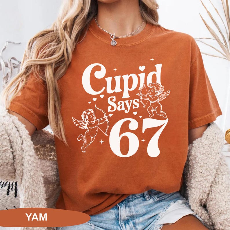 May include: Burnt orange t-shirt with the text "Cupid Says 67" in white. The design includes two cherubs with bows and arrows. The shirt is a short-sleeved, crew neck style.