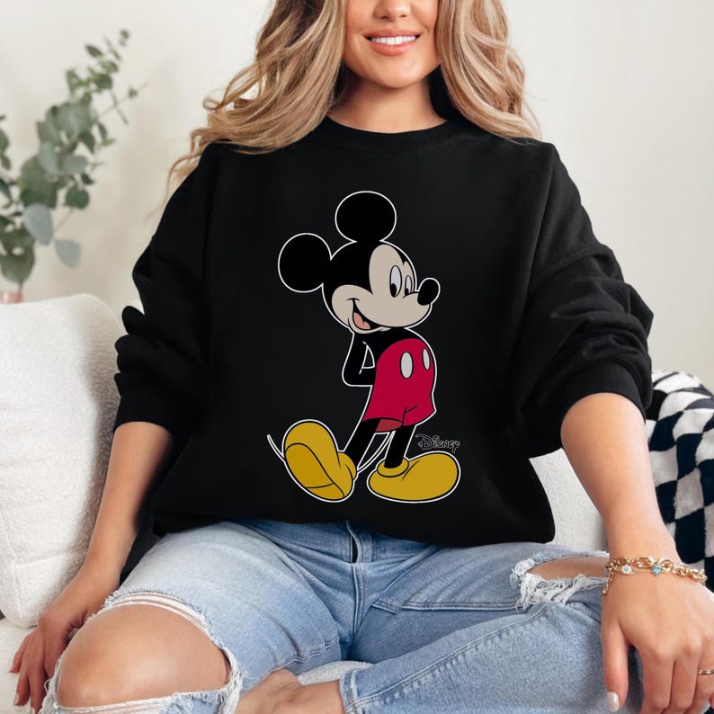 Op de afbeelding: Zwarte sweatshirt met een illustratie van Mickey Mouse. Mickey wordt afgebeeld met een wit gezicht, zwarte oren, een rode broek en gele schoenen. Het Disney-logo is zichtbaar.