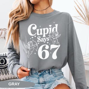 May include: Gray long-sleeve sweatshirt with the text "Cupid Says 67" in white. The design includes two cherubs with bows and arrows, and hearts. The word "GRAY" is printed at the bottom.