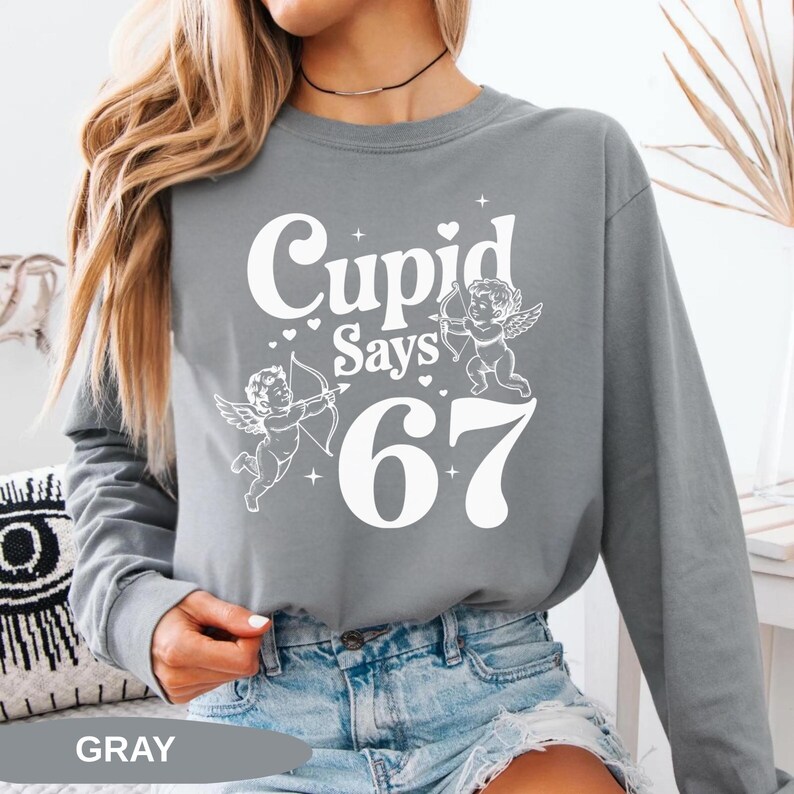 May include: Gray long-sleeve sweatshirt with the text "Cupid Says 67" in white. The design includes two cherubs with bows and arrows, and hearts. The word "GRAY" is printed at the bottom.
