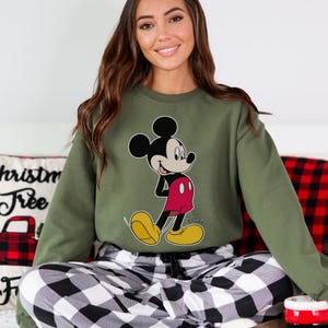 Op de afbeelding: Een olijfgroene sweatshirt met een Mickey Mouse-afbeelding in zwart, rood en geel. De sweatshirt wordt gecombineerd met een zwart-wit geruite pyjamabroek. Het Disney-logo is zichtbaar op de sweatshirt.