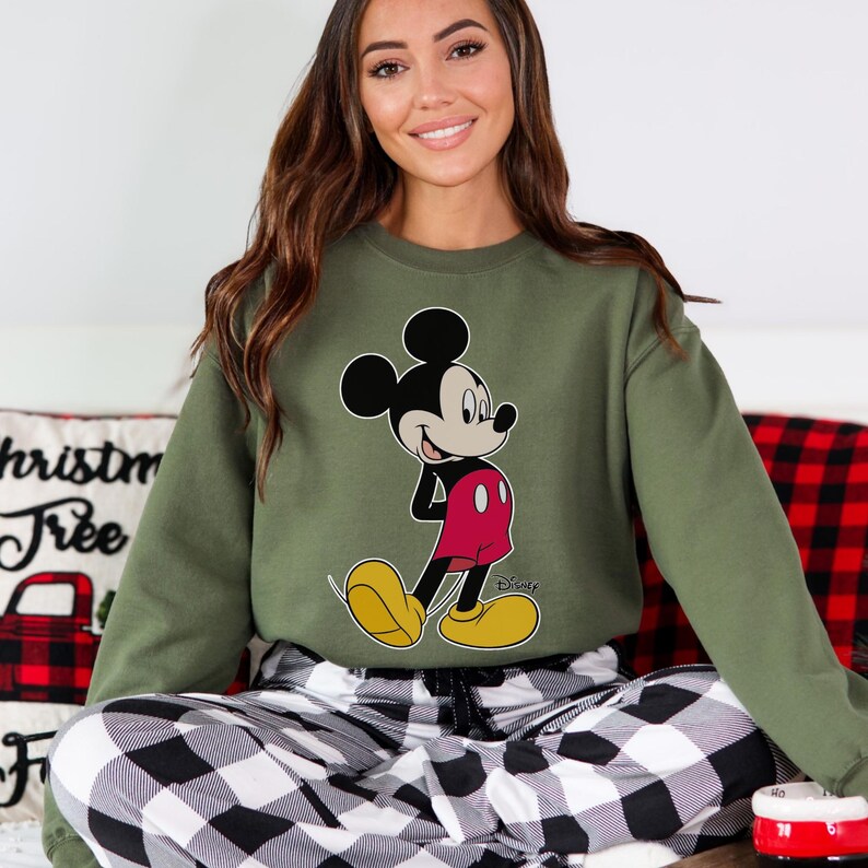 Op de afbeelding: Een olijfgroene sweatshirt met een Mickey Mouse-afbeelding in zwart, rood en geel. De sweatshirt wordt gecombineerd met een zwart-wit geruite pyjamabroek. Het Disney-logo is zichtbaar op de sweatshirt.