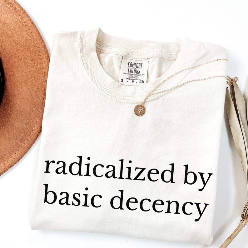 Puede incluir: Camiseta color crema con la frase "radicalized by basic decency" en negro. Un collar dorado con un colgante y un sombrero marr&oacute;n est&aacute;n junto a la camiseta. La camiseta tiene una etiqueta Comfort Colors.