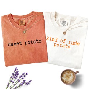 Könnte beinhalten: Zwei T-Shirts, eines in Koralle und eines in Weiß, mit den Aufdrucken "sweet potato" und "kind of rude potato". Jedes Shirt hat eine goldene Halskette. Ein Zweig Lavendel und eine Tasse Kaffee sind ebenfalls im Bild.