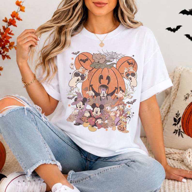 May include: A white short-sleeve t-shirt featuring a large graphic print of a central orange pumpkin with Mickey Mouse ears, surrounded by cartoon characters in Halloween costumes, ghosts, bats, and spiderwebs in shades of orange, brown, and black.