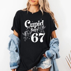 May include: Black t-shirt with the text "Cupid Says 67" in white, featuring two cherub illustrations. The cherubs are holding a bow and arrow. The shirt is worn with a denim jacket and shorts.