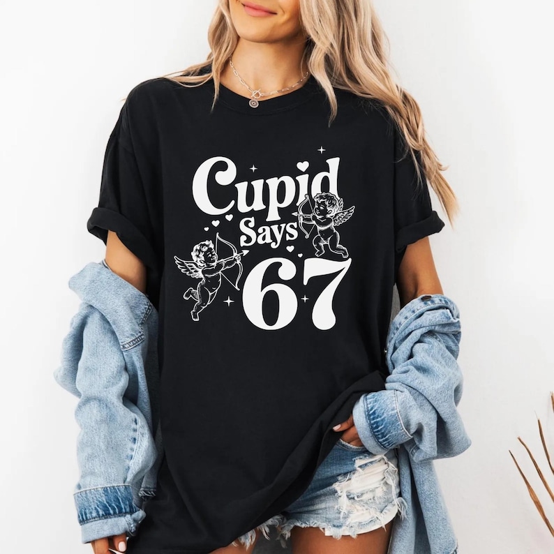May include: Black t-shirt with the text "Cupid Says 67" in white, featuring two cherub illustrations. The cherubs are holding a bow and arrow. The shirt is worn with a denim jacket and shorts.