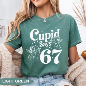 May include: Light green t-shirt with the text "Cupid Says 67" in white. The design includes two cherubs with bows and arrows, and small heart accents. The words "LIGHT GREEN" are at the bottom.