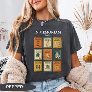 May include: Dark gray t-shirt with the text "IN MEMORIAM 2025". The shirt features postage stamp-style graphics with themes like Free Press, Climate Action, and Equality & Inclusion. The word "PEPPER" is printed at the bottom of the shirt.