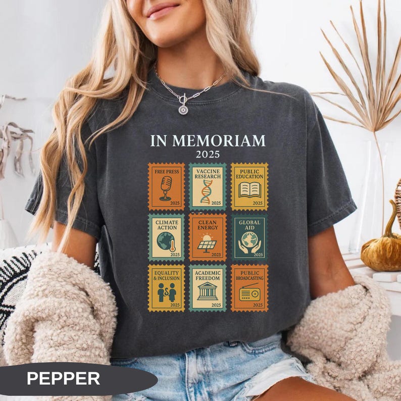 May include: Dark gray t-shirt with the text "IN MEMORIAM 2025". The shirt features postage stamp-style graphics with themes like Free Press, Climate Action, and Equality & Inclusion. The word "PEPPER" is printed at the bottom of the shirt.