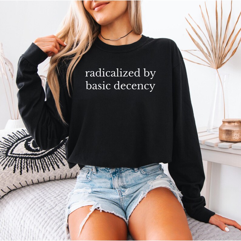 Puede incluir: Sudadera negra de manga larga con el texto blanco "radicalized by basic decency". La sudadera se combina con pantalones cortos de mezclilla. La imagen muestra un estilo casual y cotidiano.