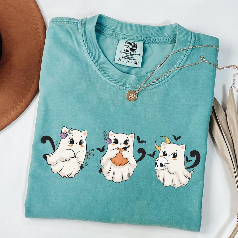 May include: Teal Comfort Colors t-shirt featuring three cartoon ghost cats. Each cat has a unique design, including flowers, a pumpkin, and a skull. A gold necklace with a pendant is draped across the shirt.