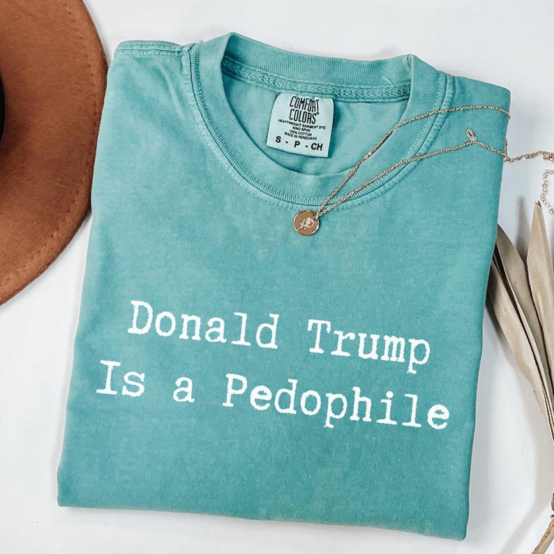 以下が含まれることがあります： 「Donald Trump Is a Pedophile」の白文字がプリントされた、ターコイズ色のComfort Colors Tシャツ。 金色のネックレスとペンダントがシャツの上に置かれています。 茶色の帽子が左上に少し見えています。
