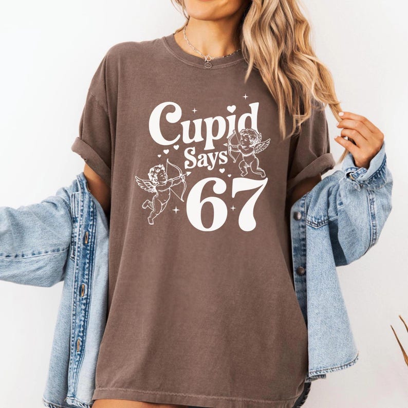 May include: A brown t-shirt with the text "Cupid Says 67" in white, featuring two cherub illustrations. The shirt has rolled-up sleeves and is worn with a denim jacket.