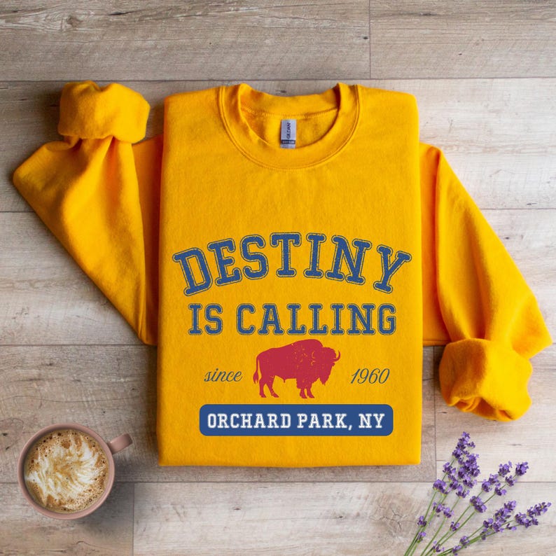 May include: A golden yellow sweatshirt with the text "DESTINY IS CALLING" in blue lettering. Below the text is a red bison graphic and the words "ORCHARD PARK, NY". The sweatshirt is folded on a wooden surface.