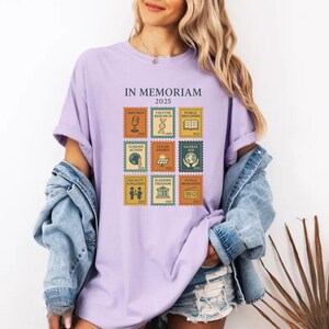 May include: A lilac-colored t-shirt with the text "IN MEMORIAM 2025" above a grid of nine postage stamp-style graphics. The graphics depict themes like "FREE PRESS," "VACCINE RESEARCH," and "PUBLIC EDUCATION." The shirt is worn with a denim jacket.
