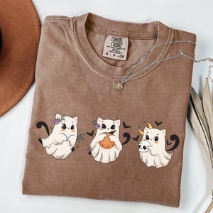 May include: Brown Comfort Colors t-shirt featuring three cartoon ghost cats with Halloween-themed accessories. The cats are white with black accents, and the shirt has a crew neck. A silver necklace with a pendant is also visible.