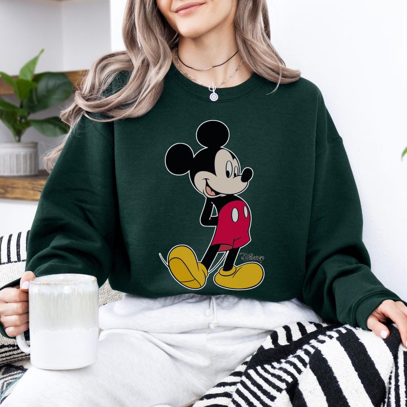 Op de afbeelding: Donkergroene sweatshirt met een Mickey Mouse-afbeelding. Het stripfiguur draagt een rode broek, gele schoenen en zwarte handschoenen. De sweatshirt heeft een ronde hals en is effen van kleur. Het Disney-logo is zichtbaar.