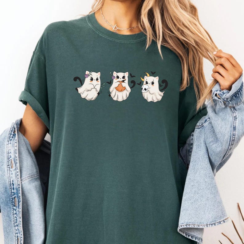 May include: Dark green t-shirt featuring three cartoon ghost cats. Each ghost cat has a unique Halloween-themed accessory: a bow, a pumpkin, and a crescent moon with bats. The shirt has short sleeves and a relaxed fit.