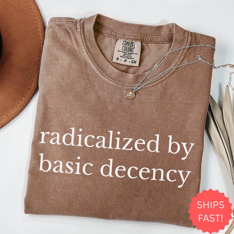 Puede incluir: Una camiseta marr&oacute;n Comfort Colors con la frase "radicalized by basic decency" impresa en blanco. Un collar plateado con un peque&ntilde;o colgante descansa sobre la camiseta. La camiseta es talla peque&ntilde;a.