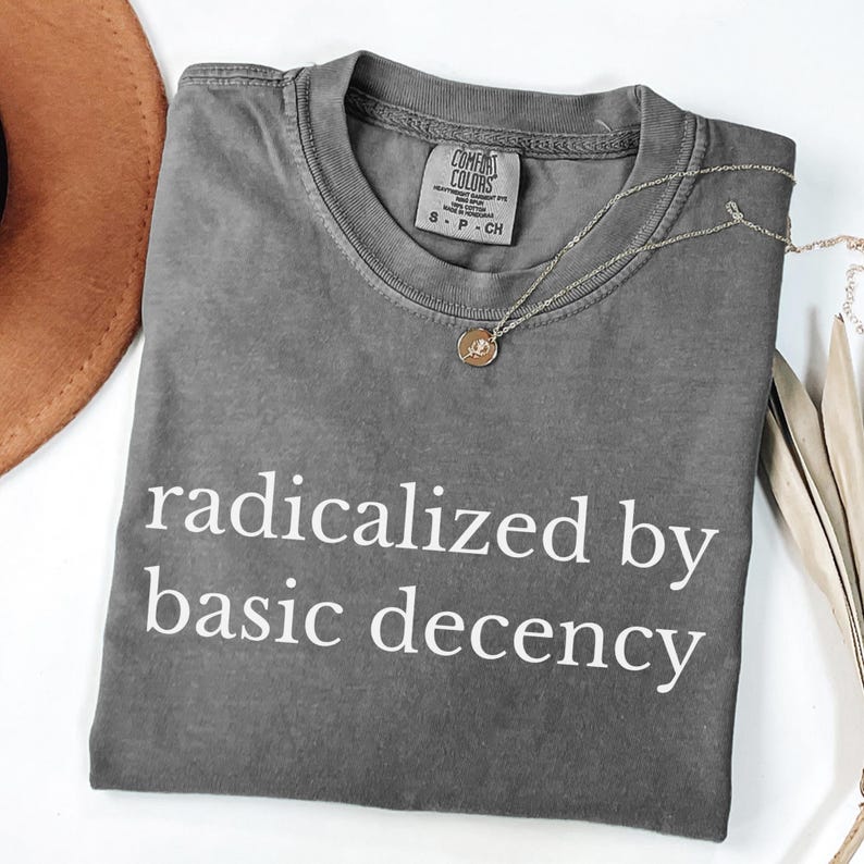Puede incluir: Una camiseta gris Comfort Colors con la frase "radicalized by basic decency" en blanco. Tambi&eacute;n se ven un collar con colgante dorado y un sombrero marr&oacute;n. La camiseta es talla S-P-CH.