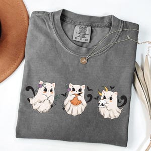 May include: A gray Comfort Colors t-shirt featuring three cartoon ghost cats with Halloween-themed accessories. The shirt has a crew neck and short sleeves. A silver necklace with a pendant is draped across the shirt.
