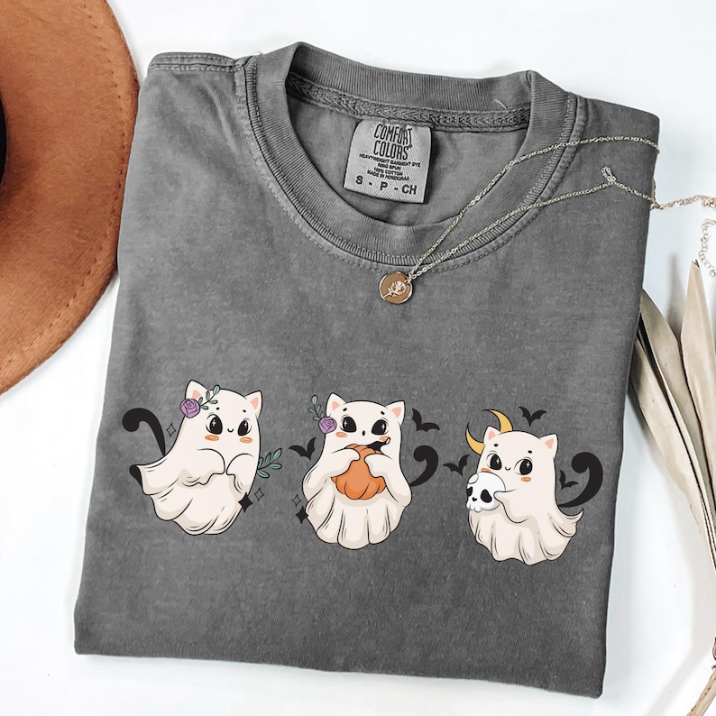 May include: A gray Comfort Colors t-shirt featuring three cartoon ghost cats with Halloween-themed accessories. The shirt has a crew neck and short sleeves. A silver necklace with a pendant is draped across the shirt.