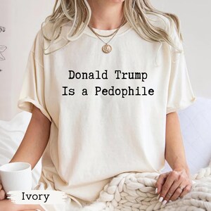 以下が含まれることがあります： 「Donald Trump Is a Pedophile」の黒い文字がプリントされたアイボリー色のTシャツ。クルーネックのシャツです。「Ivory」の文字が画像の下部に印刷されています。